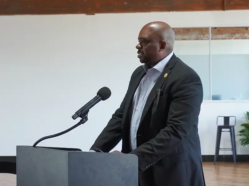Person speaking at a podium with a microphone in a modern indoor venue with exposed wooden ceiling beams and a wooden floor.