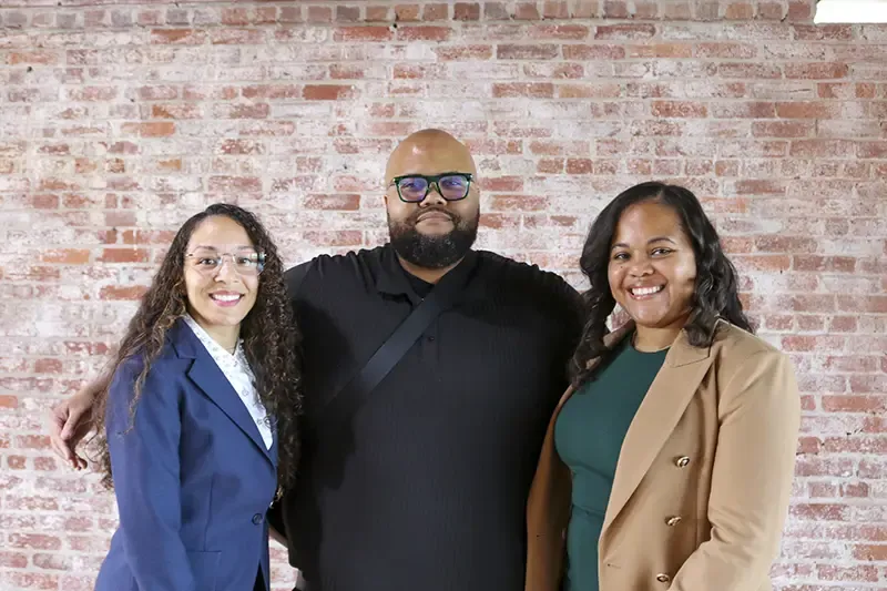 Three professionals posing together indoors against a red brick wall backdrop, dressed in business casual and formal attire, smiling at camera.