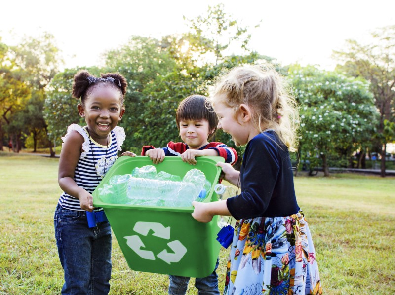Three children holding a green recycling bin filled with plastic bottles in a park