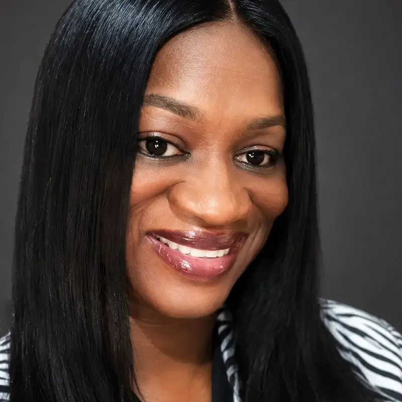 Professional headshot portrait of a person with long straight black hair, smiling warmly at the camera against a neutral gray background.