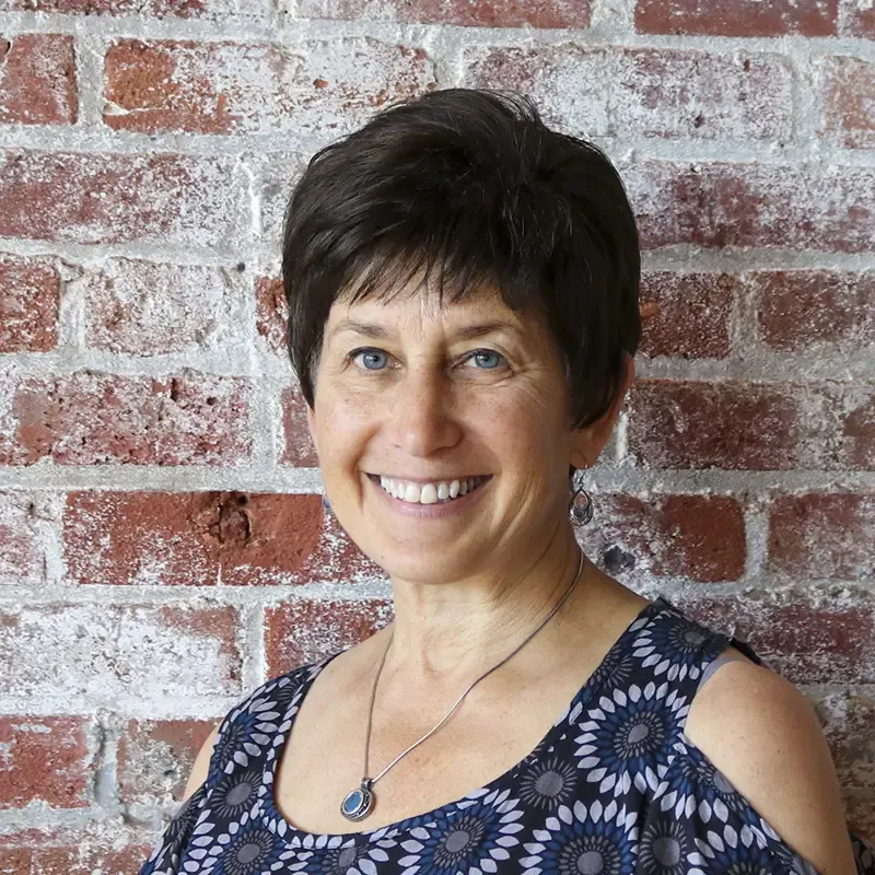 Professional headshot portrait of a person with short dark hair and warm smile, wearing a patterned navy sleeveless top and pendant necklace, posed against a brick wall.